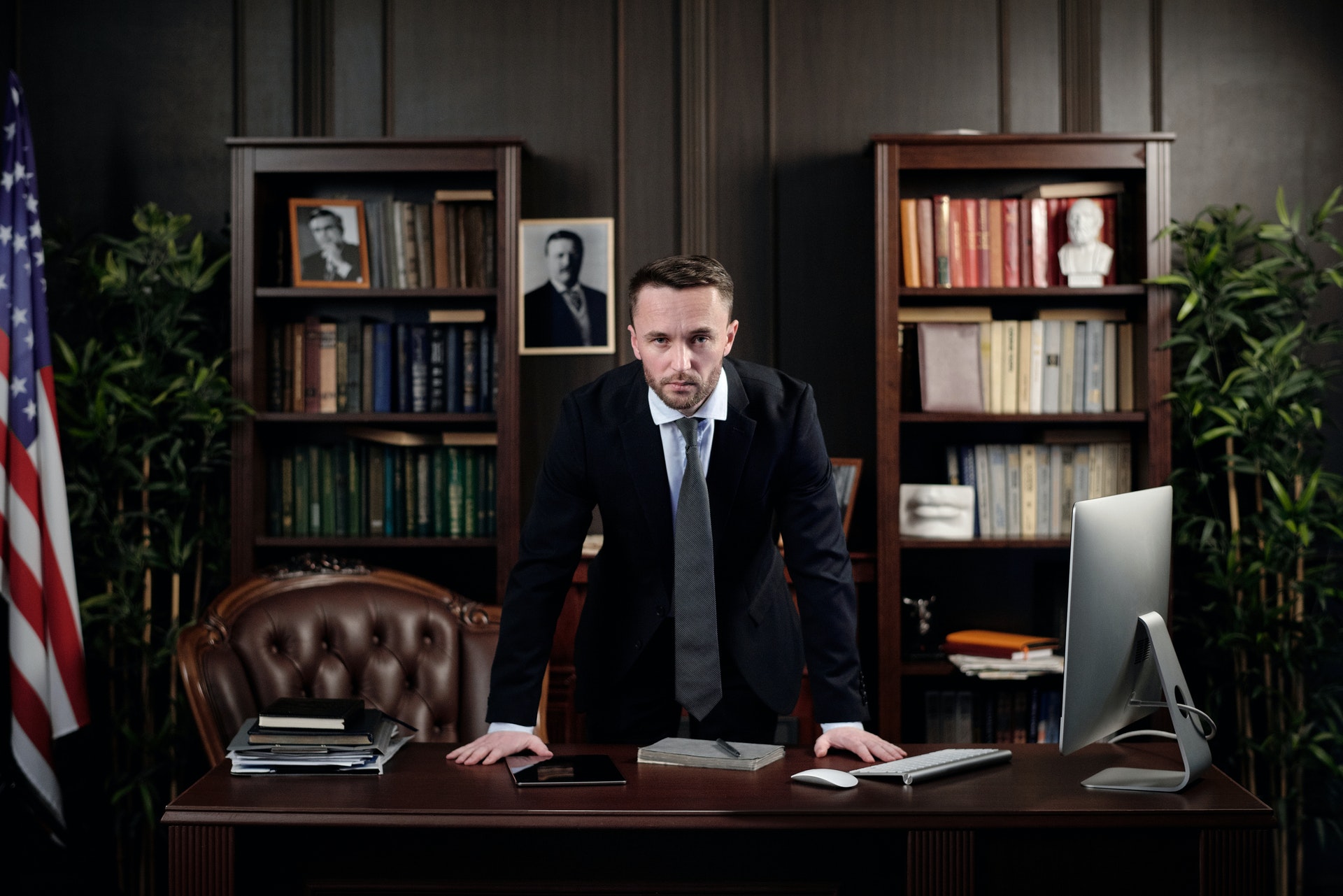 A man in a dark suit leaning on a desk in a wood-panelled office, with bookshelves and a US flag behind him