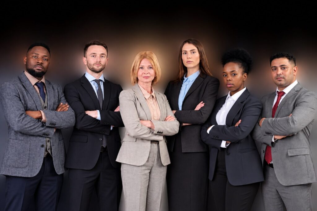 Six people in business suits standing in a row with their arms folded, against a dark background